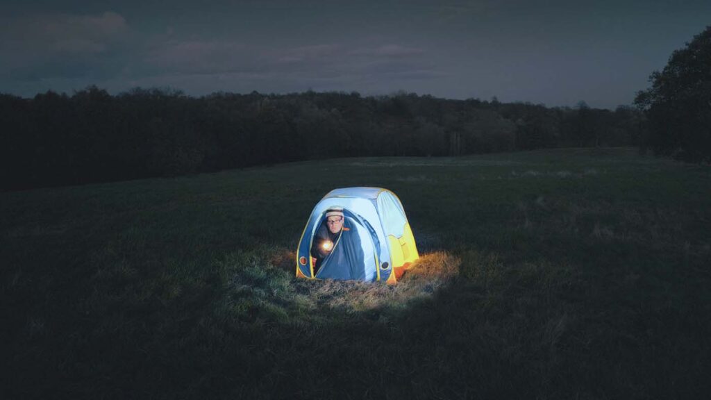 man holding a flashlight camping in small tent in the middle of an empty field